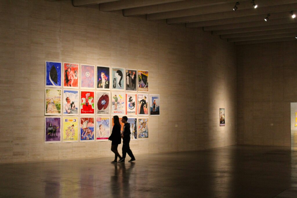 “Wide gallery space with illuminated poster artworks and visitors walking through the exhibit.”
Photo by Rui Alves on Unspashhttps://unsplash.com/photos/a-man-and-woman-standing-in-a-room-with-a-wall-of-paintings-FdOqTIgy1_s